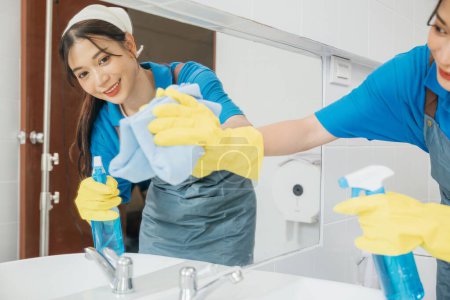 Photo for Rubber-gloved maid cheerfully wipes bathroom mirror with a rag ensuring shining reflections. Professional cleaning service highlights hygiene and modern bathroom care. Maid cleaning at home - Royalty Free Image