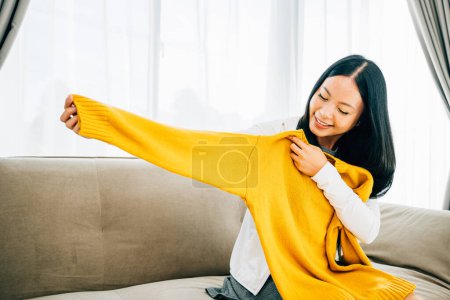 Photo for Happy Asian woman reveals a shirt from a box smiling with delight at home. Satisfied shopper unboxes online order unveiling new clothes. Delivery and home shopping concept. - Royalty Free Image
