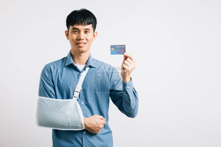Photo for A man with a damaged arm displays confidence, wearing a support splint and settling medical expenses via credit card. Happy Asian man in a sling, isolated on white, emphasizing health care solutions. - Royalty Free Image
