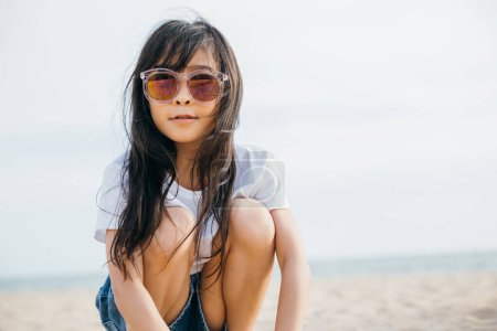 Foto de Niña de moda en gafas de sol de moda sentada en una tumbona en la playa. Retrato sonriente que emana alegría y felicidad. Cabello ventoso ropa de playa elegante y relajación sin preocupaciones - Imagen libre de derechos