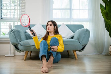 Photo for Asian young woman sitting floor using mosquito swatter or electric net racket, female killing mosquitoes she holding fly swatter like weapon in living room at home, Insect killer - Royalty Free Image