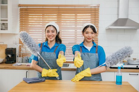 Foto de Dos mujeres de servicio de limpieza asiáticas en el mostrador de la cocina con aerosol de niebla de plumero y trapo ilustran el trabajo en equipo eficaz del hogar. Retrato limpio dos sirvienta uniforme trabajando empleado sonriente. - Imagen libre de derechos