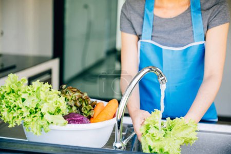 Photo for A joyful woman washes and prepares fresh vegetables in her kitchen sink for a vibrant salad enjoying her vegetarian lifestyle with healthy clean eating habits at home. - Royalty Free Image
