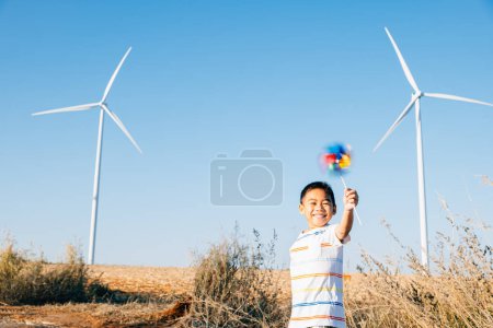 Photo for Amidst wind turbines playful boy with pinwheel toy symbolizes excitement of wind energy exploration. Showcases clean electricity innovation in a countryside windmill setting against a serene blue sky. - Royalty Free Image