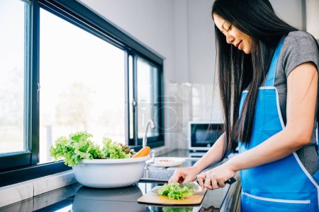 Foto de Una mujer sonriente en un delantal prepara una cena saludable cortando verduras para una deliciosa ensalada en su cocina casera. Primer plano de una alegre ama de casa haciendo una comida nutritiva. - Imagen libre de derechos