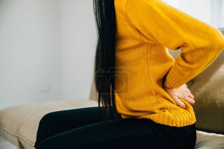 Photo for Highlighting unbearable back pain, Asian woman on sofa holds her lower back. Illustrating chronic backache discomfort and the need for medical care and attention. healthcare and problem concept - Royalty Free Image