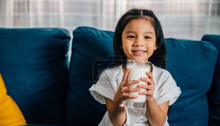 Téléchargez les photos : Asiatique enfant jouit d'un verre de lait sur le canapé leur bonheur et l'innocence brille à travers. Cette photo représente magnifiquement l'importance des soins de santé quotidiens et une bonne nutrition pour les enfants. - en image libre de droit