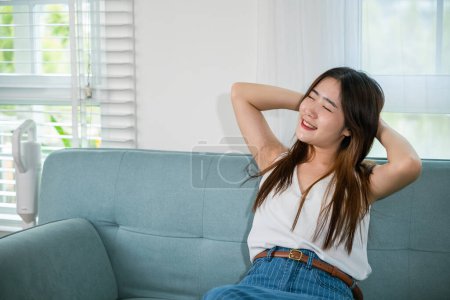 Téléchargez les photos : Asiatique jeune femme jouissant de temps libre détente mettre bras derrière la tête sur le canapé dans le salon à la maison, heureux sentiment féminin détente confort après les devoirs - en image libre de droit