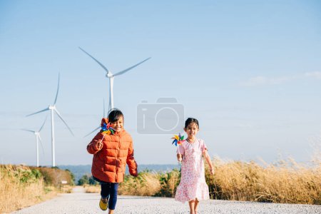 Foto de Dos niñas sosteniendo alfiler alegremente a cargo de molinos de viento. La familia cercana a las turbinas encarna la esencia de energía limpia. Niños felicidad molino de viento círculo contra el sol simboliza comunidad alegre sostenible. - Imagen libre de derechos