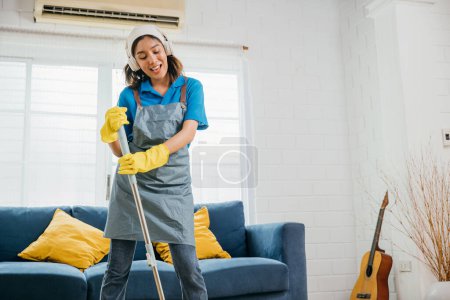 Photo for Maid musical chores, Asian teenager listens dances sings with headphones. Joyful occupation paired with music excitement. Modern cleaning with tech twist. Give me melody, Happy and Fun During Cleaning - Royalty Free Image