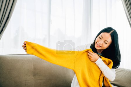 Foto de En casa una mujer asiática felizmente revela una camisa de una caja sonriendo. Comprador excitado descajas entrega en línea que muestra ropa nueva. Entrega y concepto de compras en línea. - Imagen libre de derechos