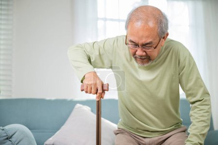Photo for Elderly man suffering from knee pain ache holding handle of cane, senior old man typing to stand up from sofa alone with walking cane stick to walk at home, retirement medical healthcare concept - Royalty Free Image