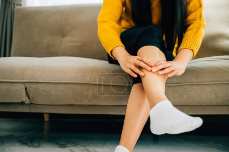 Photo for A woman on sofa holds her injured ankle showcasing pain. Depicting health care varicose vein prevention and emphasizing leg recovery and pain relief concept. medical - Royalty Free Image