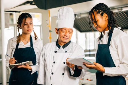 Téléchargez les photos : Chef asiatique senior éduque les étudiants multiraciaux dans une cuisine de restaurant. Mettre l'accent sur l'apprentissage du travail d'équipe et la prise de notes. Formation professionnelle en cours. Edocation alimentaire - en image libre de droit