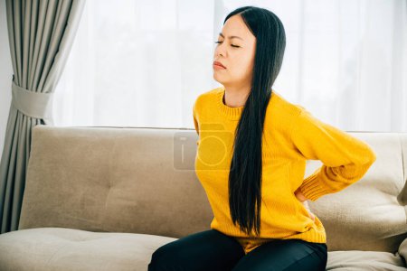 Photo for Highlighting backache, Asian woman seated experiencing unbearable lower back pain. Illustrating chronic back pain discomfort and the importance of medical care for back problems. - Royalty Free Image
