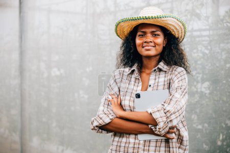 Photo for Greenhouse shop owner a successful young woman in a checked shirt and apron stands with arms crossed smiling. A black woman holding a tablet poses confidently in front of the greenhouse. - Royalty Free Image