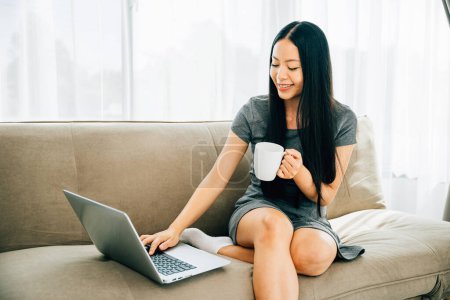 Relaxed woman on sofa sips coffee watches laptop education. Engaged in learning relaxation in cozy living room. Enjoying remote education and comfort. Diverse education experiences.