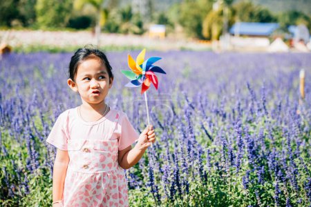 Foto de Retrato de una niña sonriente en un jardín de flores sosteniendo un molinete de juguete. La primavera trae alegría y el molinete giratorio significa diversión infantil libertad y felicidad en naturalezas ambiente brillante y soleado. - Imagen libre de derechos