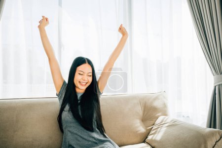 Photo for Happy woman on sofa raises arms stretches after sitting for long time. Lifestyle of a cheerful businesswoman in a luxury living room. Wellbeing joy and carefree relaxation. - Royalty Free Image