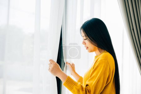 Téléchargez les photos : Une jeune femme se réveille tôt en ouvrant les rideaux en souriant à la vue sereine se sentant rafraîchi et détendu à la maison. Faire place à la joie du matin détente et un début de journée gai. - en image libre de droit