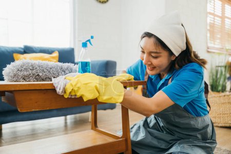 Téléchargez les photos : Une femme au foyer joyeuse portant des gants jaunes nettoie la table dans son salon avec soin. Son dévouement à la propreté et à l'hygiène à la maison est évident dans son programme de nettoyage de routine. pucelle bureau propre. - en image libre de droit