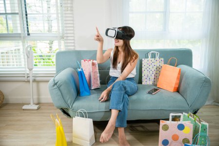 Téléchargez les photos : Heureuse jeune cliente portant des lunettes VR 3D casque de réalité virtuelle avec des sacs à provisions autour d'elle toucher et pointant sur l'air pour faire du shopping en ligne dans le salon à la maison, cyberespace numérique - en image libre de droit