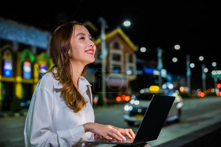 Foto de En una bulliciosa calle de la ciudad por la noche, una mujer trabaja duro en su computadora portátil, utilizando la comodidad y la movilidad de su dispositivo para mantenerse conectada en la era digital. - Imagen libre de derechos