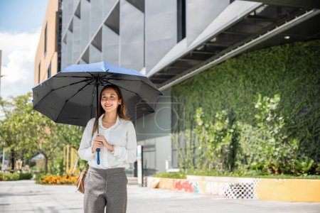 Foto de Bajo el sol caliente, una joven empresaria asiática camina al trabajo sosteniendo un paraguas. Su expresión severa y su atuendo profesional reflejan su determinación y éxito en el mundo de los negocios. - Imagen libre de derechos