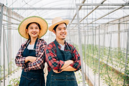 Foto de Pareja asiática agricultores prosperan en la granja hidropónica de tomate. Marido sonriente y esposa cruzaron los brazos llevando verduras de calidad. Retrato de éxito en el invernadero. Agricultura felicidad. - Imagen libre de derechos
