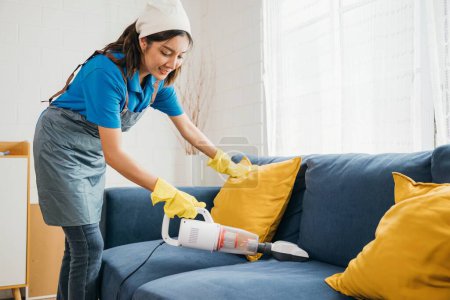 Photo for In a cozy setting an Asian woman focuses on housework using a vacuum machine cleaner to clean a sofa. Her dedication to hygiene and furniture care exemplifies a modern approach to cleaning. - Royalty Free Image