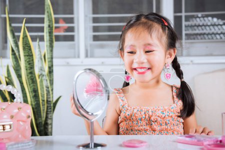 Foto de Niño feliz es hermoso maquillaje con juguete de cosméticos, asiática adorable niña divertida haciendo maquillaje su cara que se mira en el espejo y la aplicación de lápiz labial rojo a la boca, actividad de aprendizaje para ser mujer - Imagen libre de derechos