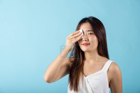 Téléchargez les photos : Facial Beauty Treatment Hygiene skin. Asian beautiful young woman removing cosmetic make up using pad cotton face cleaner disc, studio shot isolated on blue background, Happy female cleaning eyes - en image libre de droit