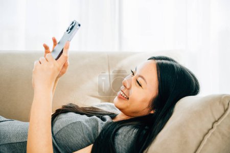 Téléchargez les photos : Femme asiatique sur canapé textes sur smartphone souriant. Bénéficiant de détente bavardage et achats en ligne. Technologie moderne pour la communication et la connexion à la maison. - en image libre de droit