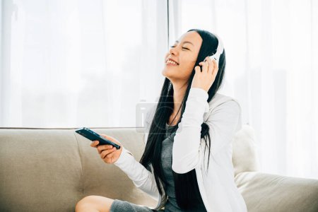 Photo for Young woman on sofa enjoys leisure time listens to music using smartphone and headphones. Embracing relaxation enjoyment and modern technology at home. - Royalty Free Image