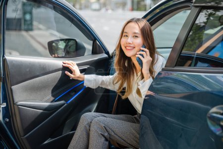 Foto de Una mujer de negocios asiática, sentada en su coche de lujo, sonríe mientras consigue un nuevo contrato por teléfono. Su puerta de coche abierta simboliza la puerta al éxito empresarial en la ciudad moderna. - Imagen libre de derechos