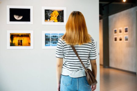 Photo for Asian woman standing she looking art gallery in front of colorful framed paintings pictures on white wall, young female watch at photo frame to leaning against at show exhibition gallery, Back view - Royalty Free Image