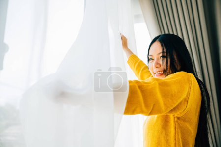 Foto de Ilustrando la alegría de la mañana, Mujer abre cortinas sonrisas en la vista refrescante sensación de felicidad y relajación en casa. Abrazar un comienzo alegre y despreocupado del día. - Imagen libre de derechos