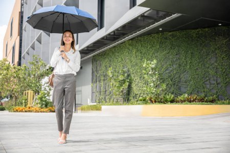 Foto de Caminando a la oficina en un día caluroso, una joven empresaria sostiene un paraguas para protegerse de los rayos del sol. Su comportamiento profesional y maquillaje reflejan su determinación y éxito. - Imagen libre de derechos