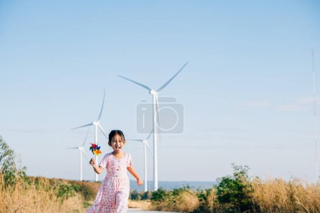 Foto de Niñas interacción juguetona cerca de molinos de viento sosteniendo molinos mientras se ejecuta. Adoptar la educación en energía eólica promoviendo la electricidad limpia y una infancia alegre. - Imagen libre de derechos
