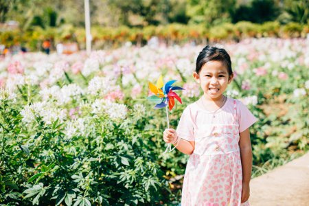 Foto de Retrato de una niña alegre en un jardín de flores sosteniendo un molinete de juguete. La animada temporada de primavera evoca felicidad y el molinete giratorio encarna la alegría y la libertad de la infancia en la belleza soleada naturalezas. - Imagen libre de derechos