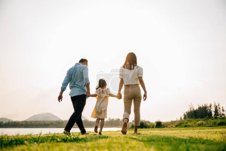 Foto de Happy Asian family walking and playing together in a beautiful nature setting, with green grass and a sunny background, on a summer day, Family day, back view - Imagen libre de derechos