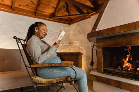 Photo for Senior African woman reading book while warming in front of fireplace in her house - Royalty Free Image