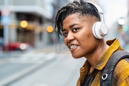 Photo for African woman listening to music with wireless headphones while waiting for public transportation in the city street - Royalty Free Image