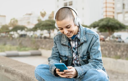 Foto de Chica cabeza afeitada usando teléfono inteligente móvil mientras escucha música con auriculares en la calle de la ciudad - Imagen libre de derechos