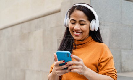 Photo for Happy southeast asian woman listening to music with mobile smartphone and headphones in the city center - Royalty Free Image