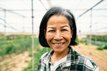 Téléchargez les photos : Heureuse femme d'Asie du Sud-Est souriant à la caméra tout en travaillant à l'intérieur de la serre agricole - Concept de style de vie des agriculteurs - en image libre de droit