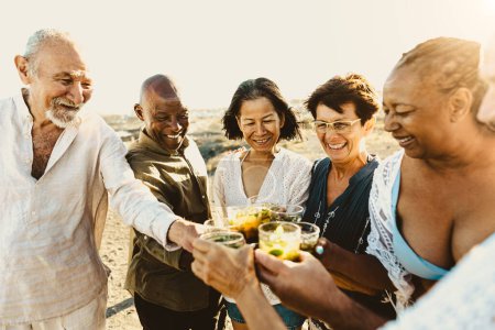 Téléchargez les photos : Heureux amis aînés multiraciaux s'amuser à boire et à griller des mojitos sur la plage pendant l'heure du coucher du soleil - Les personnes âgées profitant des vacances d'été - en image libre de droit