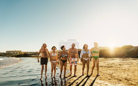 Téléchargez les photos : Heureux amis aînés multiraciaux qui s'amusent à marcher sur la plage au coucher du soleil pendant les vacances d'été - Diverses personnes âgées profitant de vacances - en image libre de droit
