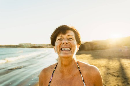 Téléchargez les photos : Heureuse femme âgée caucasienne s'amuser en souriant à la caméra sur la plage pendant les vacances d'été concept de style de vie des personnes âgées - en image libre de droit