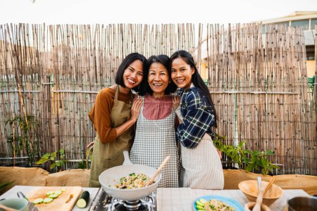 Téléchargez les photos : Heureuse famille d'Asie du Sud-Est avoir du plaisir à sourire devant la caméra tout en préparant la recette de nourriture thaïlandaise ensemble à la maison patio - en image libre de droit
